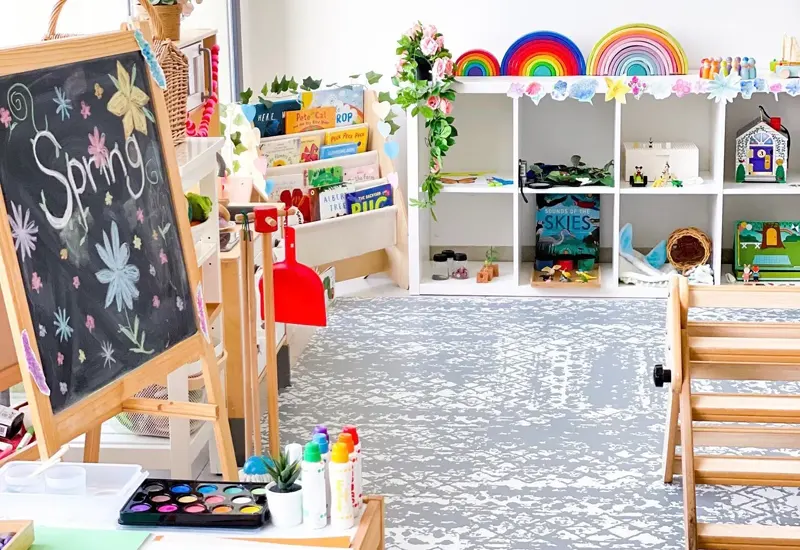 A bright and colorful classroom with a chalkboard displaying "Spring," shelves filled with books and educational toys, art supplies on a table, and decorations creating a cheerful atmosphere.