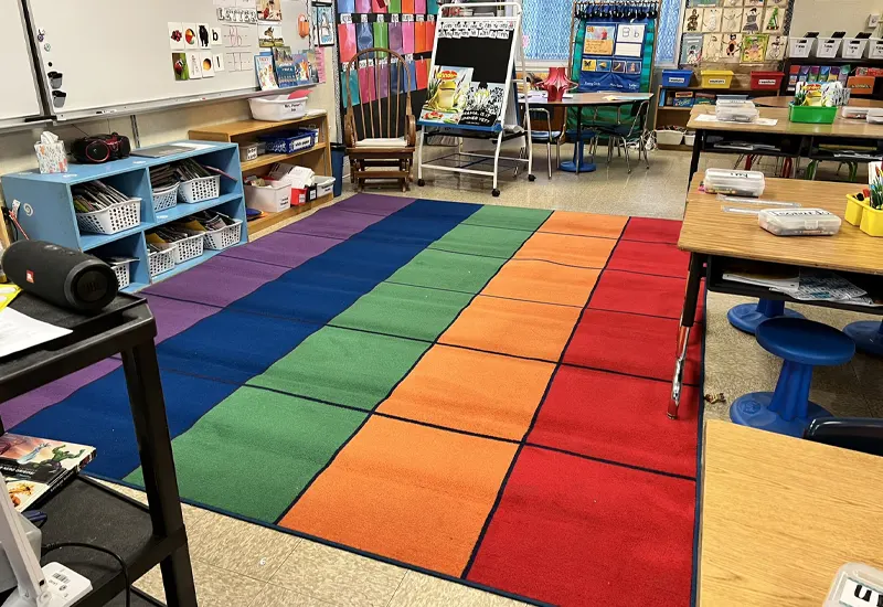 A colorful classroom with a large, multi-colored carpet in the center, surrounded by desks, chairs, and various educational materials and decorations on the walls. 