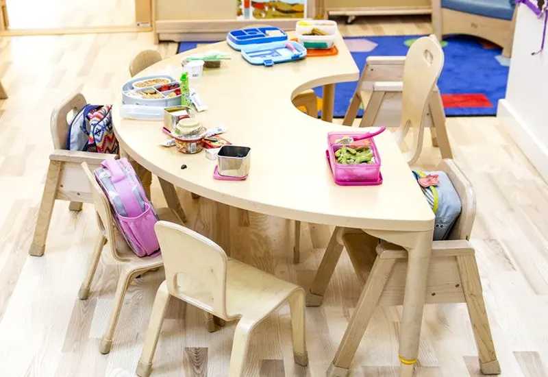 A semi-circular wooden table with children's lunchboxes and snacks, surrounded by small chairs in a bright classroom.