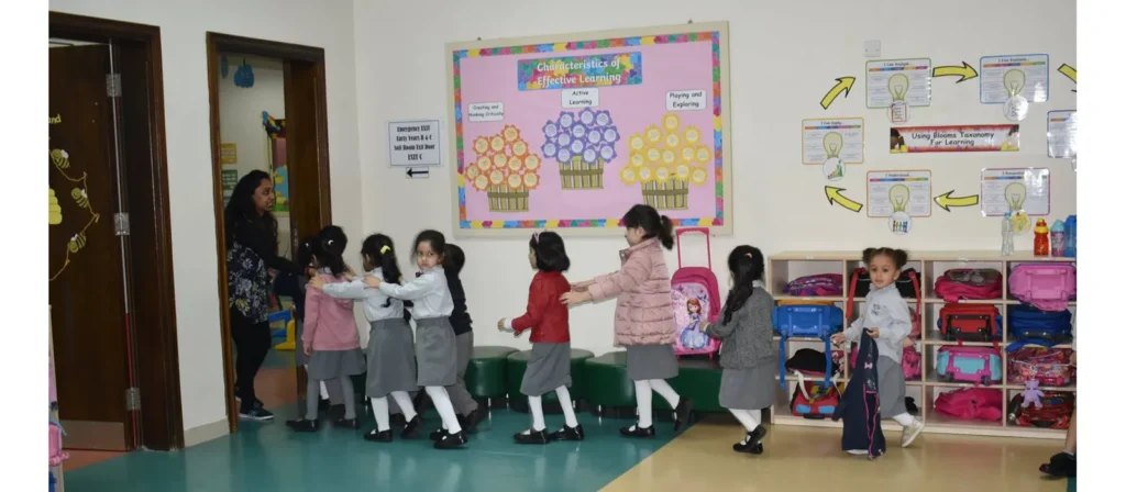 The image shows young children in a classroom, lined up and holding onto each other, being guided by a teacher towards an emergency exit. A sign indicates the location of the emergency exit, EXIT C.