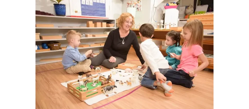 Montessori Caregiver is sitting on the floor with four children in a classroom. They are engaging with various educational toys and materials, including animal figurines and a small farm setup. The children are interacting with the toys and the teacher, who appears to be guiding or participating in their activity.
