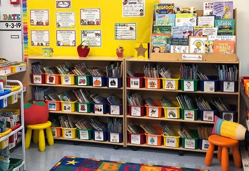The image shows a well-organized classroom storage area. Books are neatly arranged in bins on shelves, each bin labeled with a picture for easy identification. A yellow bulletin board with reading activities is displayed above the shelves. 