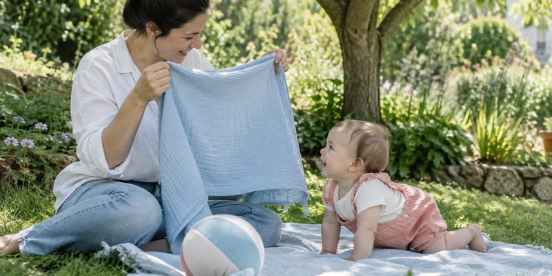 Object Permanence: What Babies Learn And How Montessori Activities Support It 1 Caregiver Playing A Gentle Hide-And-Reveal Game With An Infant Outdoors On A Pale Blue Blanket